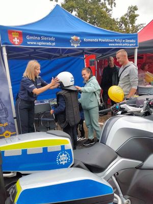 pod niebieskim namiotem stoisko policyjne na Jarmarku Powiatowym, przed namiotem stoi dziecko, któremu umundurowana policjantka zakłada elementy wyposażenia policyjnego