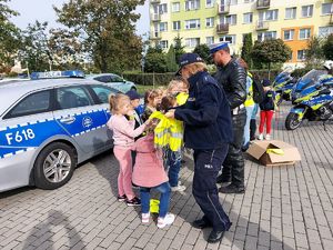 umundurowanych policjantka rozdaje dzieciom kamizelki odblaskowe, obok  policyjny radiowóz