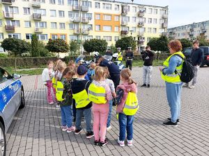 umundurowanych policjantka  i policjant rozdaje dzieciom kamizelki odblaskowe, obok  policyjny radiowóz