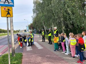 dzieci z założonymi worka mi odblaskowymi stoją przed przejściem dla pieszych razem z nimi są umundurowani policjanci