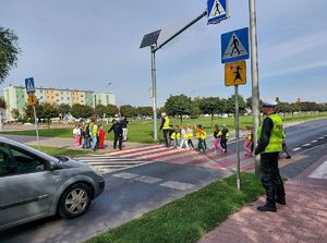 dzieci z założonymi worka mi odblaskowymi wspólnie z umundurowaną policjantka idą przez przejście dla pieszych