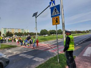 dzieci z założonymi worka mi odblaskowymi wspólnie z umundurowaną policjantka idą przez przejście dla pieszych