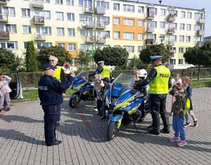 dzieci wsiadają na policyjny motocykl obok stoi policjant w kamizelce odblaskowej