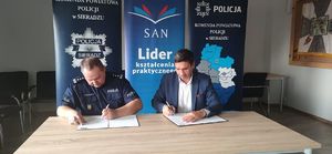 na tle bannerów Społecznej Akademii Nauk i Komendy Powiatowej Policji w Sieradzu przy stole siedzi umundurowany policjant oraz  mężczyzna, obydwaj podpisują dokument