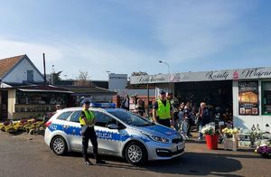 umundurowani policjanci stoją przed oznakowanym radiowozem wzdłuż parkanu cmentarza gdzie stoją kwiaty