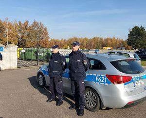 umundurowani policjanci stoją przed oznakowanym radiowozem wzdłuż parkanu cmentarza