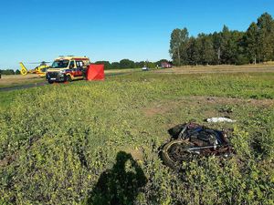 Miejsce wypadku widać drogę , samochody oznakowane służb ratunkowych, helikopter, na trawie motocykl.