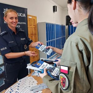 Stoisko promocyjne, przy którym stoi umundurowana policjantka, przed nią na stolikach leżą ulotki i materiały promocyjne, obok stoją dwie uczennice w mundurach, którym rozdaje ulotki.