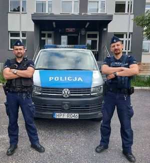 Przed budynkiem komendy przed radiowozem stoi dwóch umundurowanych policjantów.