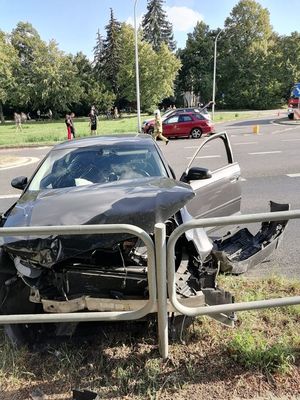 Na zdjęciu widać pojazd koloru grafitowego, który posiada uszkodzony przód pojazdu stojący  przodem do barierki. W tle na jezdni widać stojące na jezdni dwa pojazdy ułożone bokami do siebie oraz ludzi stojących na jezdni oraz trawie.