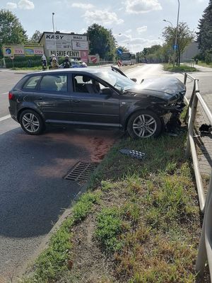 Na zdjęciu widać pojazd koloru grafitowego, stojący w poprzek jezdni i skierowany w kierunku bariery. Pojazd posiada widoczny uszkodzony przód pojazdu. Za pojazdem w tle widać stojący radiowóz oraz strażaków.