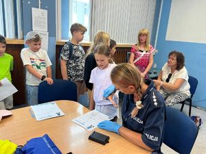 Na zdjęciu widoczna funkcjonariusza policji, która siedząc przy stole trzyma w rękach dłoń dziecka i kierują ją w stronę kartki znajdującej się na blacie stołu. W tle tuz za policjantką widać kobiety i dzieci.