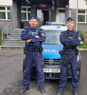 Przed budynkiem komendy na tle radiowozu stoją dwaj umundurowani policjanci.