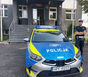 Przed budynkiem komendy przy radiowozie stoi umundurowany policjant.