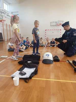 Na sali gimnastycznej szkoły umundurowany policjant prowadzi prelekcje dla dzieci.