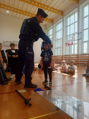 Na sali gimnastycznej szkoły umundurowany policjant prowadzi prelekcje dla dzieci.