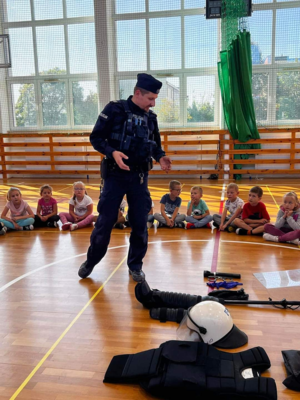 Na sali gimnastycznej szkoły umundurowany policjant prowadzi prelekcje dla dzieci.