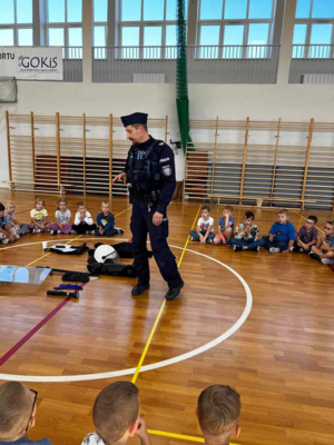 Na sali gimnastycznej szkoły umundurowany policjant prowadzi prelekcje dla dzieci.