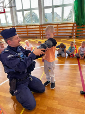 Na sali gimnastycznej szkoły umundurowany policjant prowadzi prelekcje dla dzieci.