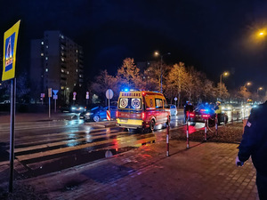 Miejsce potrącenia pieszej, widać pojazdy służb ratunkowych, umundurowanego policjanta, osoby, samochody stojącego na pasie drogowym.