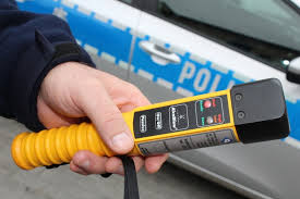Na tle drzwi oznakowanego radiowozu  dłoń z alkomatem.