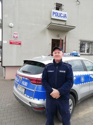 Przed budynkiem komisariatu na tle radiowozu stoi umundurowany policjant.