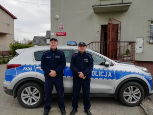 Przed budynkiem komisariatu na tle radiowozu stoją umundurowani policjanci.