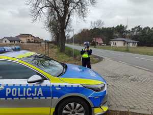 Na poboczu drogi stoi radiowóz policyjny radiowóz, umundurowana policjantka mierzy prędkość laserowym miernikiem prędkości.