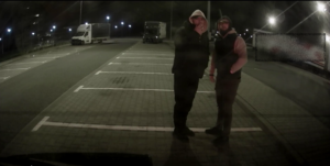 Na parkingu w porze nocnej stoją dwaj mężczyźni, którzy mają zarost na twarzy i założone kaptury na głowę.