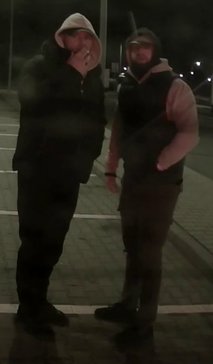 Na parkingu w porze nocnej stoją dwaj mężczyźni, którzy mają zarost na twarzy i założone kaptury na głowę.