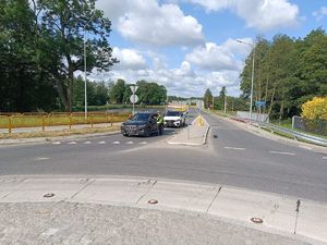 Na pasie wjazdowym na rondzie stoją dwa samochodu osobowe, przy pierwszym z nich stoi umundurowany policjant , który bada stan trzeźwości kierowcy.