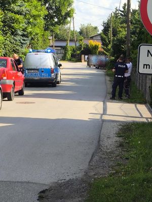 Na poboczu drogi stoją dwa policyjne radiowozy między nimi czerwony osobowy samochód, policjanci rozmawiają z kilkoma osobami.