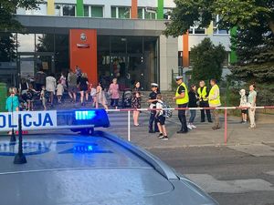 Na zdjęciu widoczny jest budynek szkolny, przed którym  na chodniku stoją dzieci policjanci wraz z dziećmi. W leym dolnym rogu widać światła z policyjnego radiowozu.