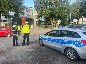 Na zdjęciu widać radiowóz policyjny, przy którym stoją policjanci. W tle widać jedną policjantkę przechodzącą przez jezdnię w kierunku budynku.