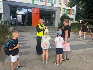 Na zdjęciu widać policjantkę stojącą na chodniku przed budynkiem szkoły, która stoi wraz z dziećmi.