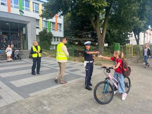 Na zdjęciu widać stojących na chodniku przed budynkiem policjantów oraz mężczyznę ubranego w żółtą odblaskową kamizelkę,  Policjantka wręcza dziewczynce na rowerze odblask.