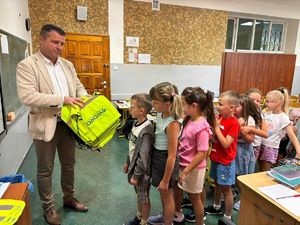 Na zdjęciu widać mężczyznę stojącego w pomieszczeniu, który trzyma w dłoniach żółte worki. Po przeciwnej stronie mężczyzny stoją dzieci.