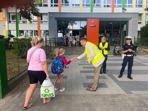 Na zdjęciu widać mężczyznę ubranego w żółtą kamizelkę, stojącego przed budynkiem szkoły. Na zdjęciu widać jak przekazuje dziecku odblask.