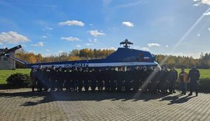 Na zdjęciu widać stojącą  na placu młodzież, wraz z opiekunami oraz policjantami.  Tuż za osobami widać stojący  niebieski helikopter.