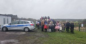 Na zdjęciu widać dzieci i młodzież, kobiety oraz trzech umundurowanych funkcjonariuszy policji z psem stojących na dworze przy radiowozie policyjnym.