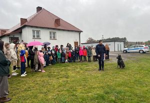 Na zdjęciu widać funkcjonariusza policji wraz psem, który wykonuje pokaz dla uczniów na terenie boiska.