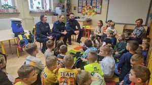 Na zdjęciu widać trzech policjantów rozmawiających z dziećmi, które siedzą na dywanie w klasie.