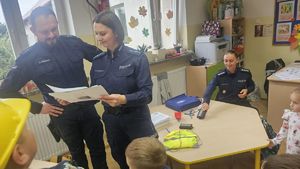 Na zdjęciu widać policjanta oraz policjantkę stojących w klasie, oglądających wykonane prace plastyczne dzieci.