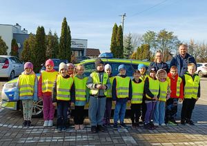 Na zdjęciu znajduję się grupa dzieci stojąca przed radiowozem policyjnym wraz z policjantami. Wszystkie dzieci na zdjęciu ubrane są w kamizelki odblaskowe.