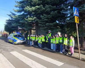 Na zdjęciu widać dzieci wraz z policjantem stojące przy krawędzi chodnika, które stoją przed przejściem dla pieszych.