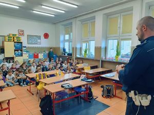 Na zdjęciu widać policjanta który skierowany jest twarzą w drugą część sali szkolnej, gdzie znajdują się dzieci,