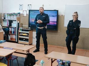 Na zdjęciu znajduję się policjantka wraz z policjantem, którzy znajdują się w sali szkolnej.