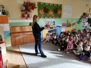 Na zdjęci widać policjantkę, która stoi w sali szkolnej i skierowana jest twarzą do grupy dzieci siedzących na dywanie.