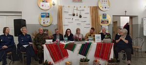 Na zdjęciu widać siedzące na krzesłach sali dwie umundurowane policjantki, obok których przed stołem siedzą mężczyzna ubrany w mundur wojskowy oraz  jednej mężczyzna ubrany w mundur strażacki, a także dwóch mężczyzn i cztery kobiety.