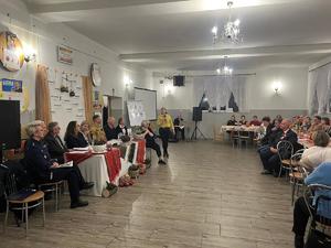 Na zdjęciu z prawej stronu widać  w pomieszczeniu osoby, które siedzą przy znajdujących z prawej stronu  stołach. Z lewej strony zdjęcia widać siedzące przy stole osoby w tym policjantkę, strażaka oraz cztery kobiety i jednego mężczyznę przy których stoi kobieta trzymająca w dłoniach mikrofon.